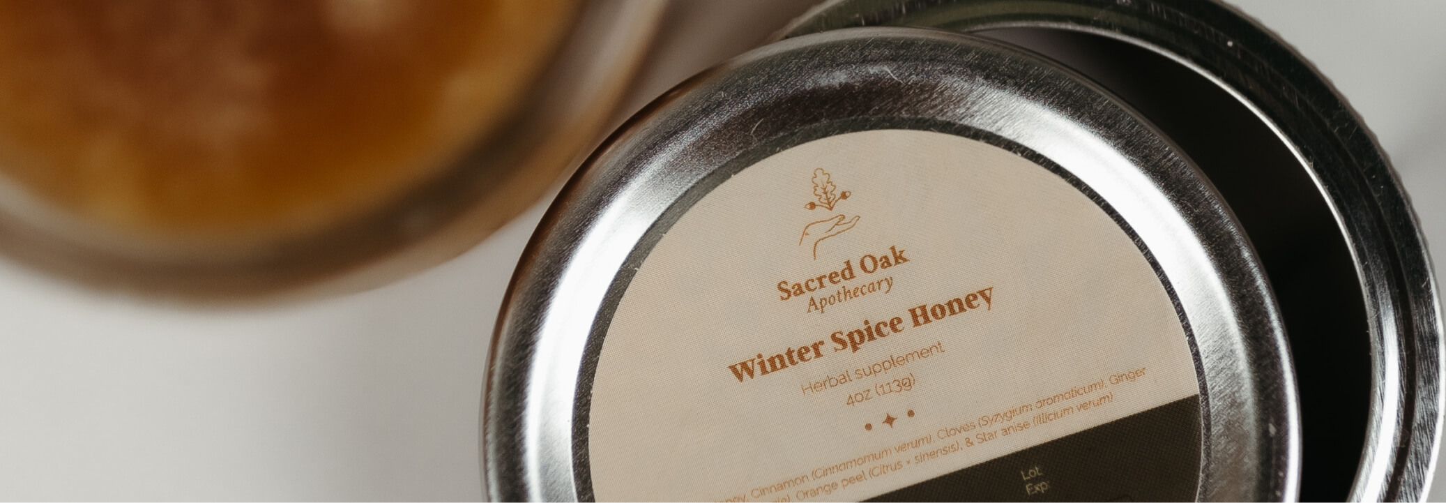 Close-up of a metal jar lid labeled ‘Sacred Oak Apothecary Winter Spice Honey’ herbal supplement