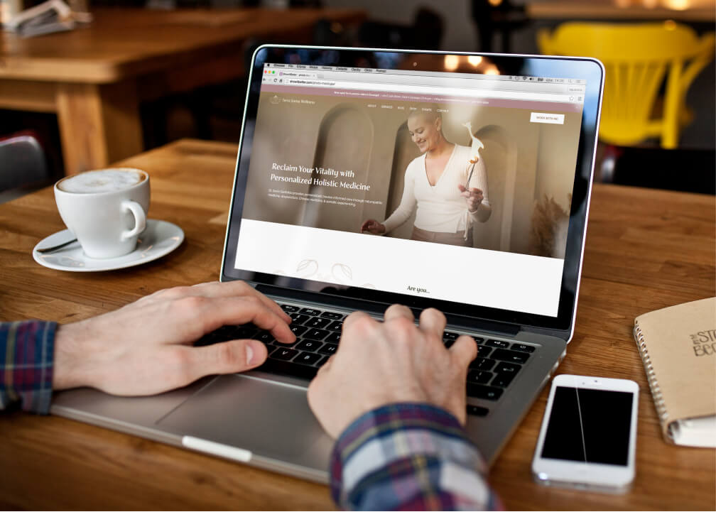 Hands typing on a laptop displaying a website headline ‘Reclaim Your Vitality with Personalized Holistic Medicine’ next to a cup of coffee
